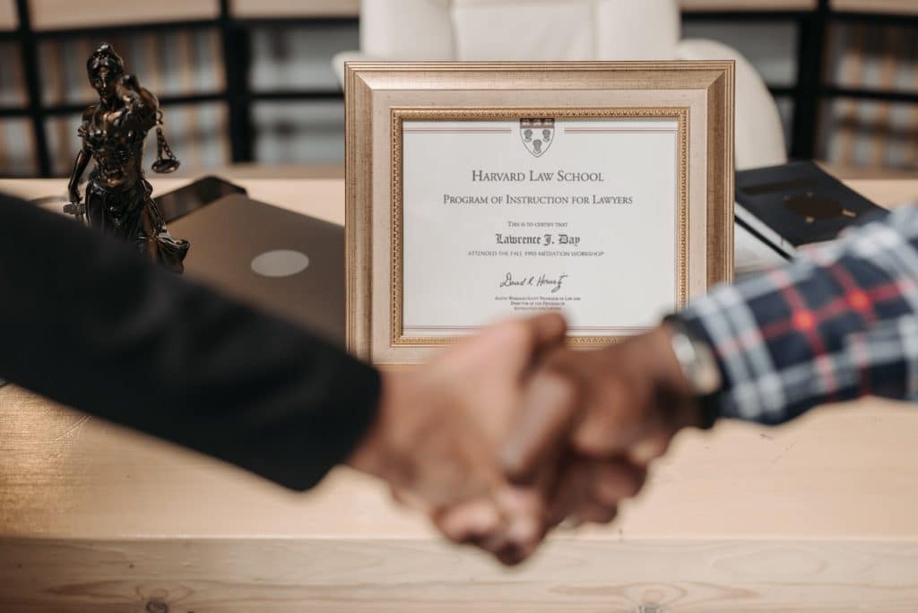 Two men shaking hands with a degree from Harvard Law School in the background. 